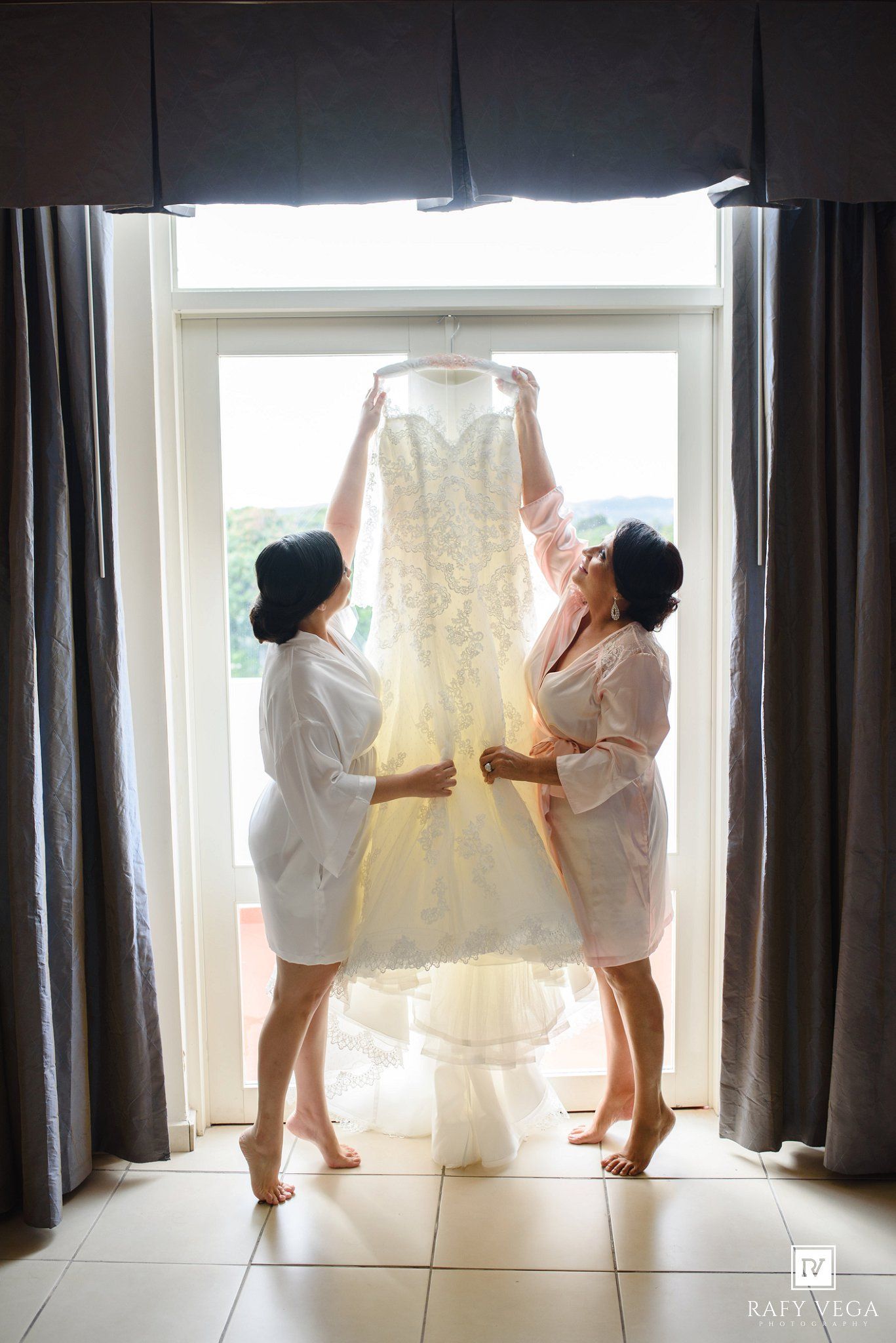 Two bridesmaids in robes hold up a lace wedding dress in front of a bright window before the ceremony.