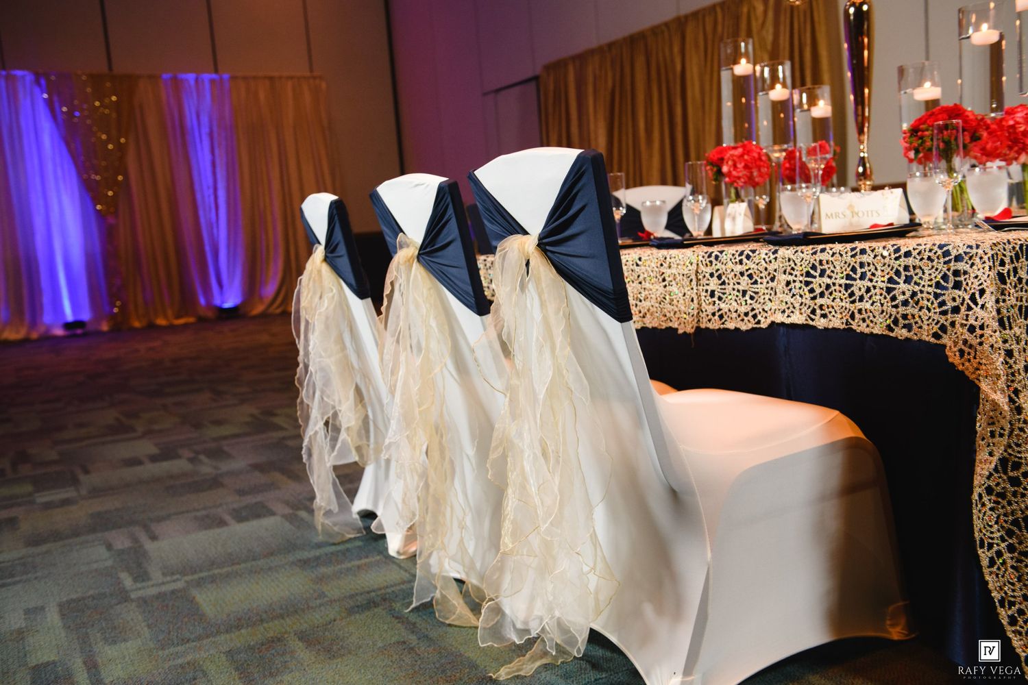 Elegant wedding reception head table with ivory ruffled chair covers, navy sashes, and red floral centerpieces in a decorated ballroom.