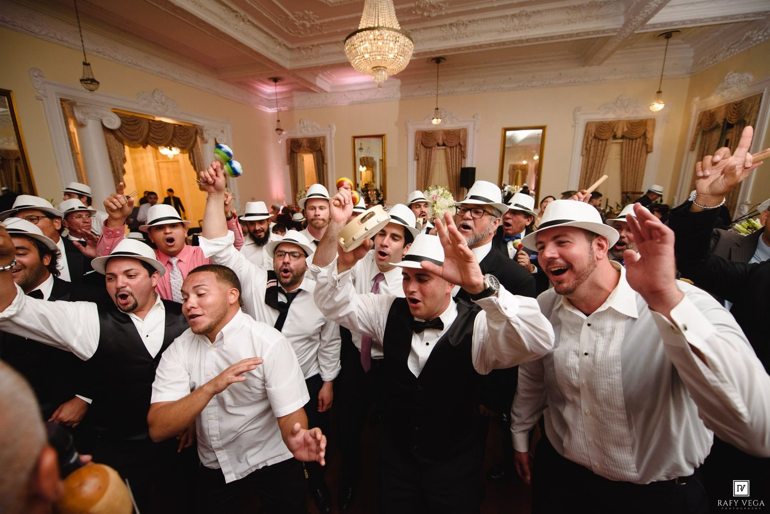 Large group of men in white fedoras and tuxedos celebrating energetically in an elegant ballroom.