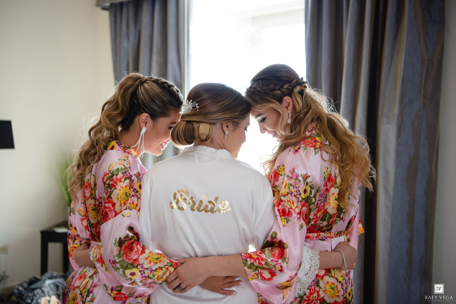 Bride in white robe with bridesmaids in floral robes sharing a tender moment before the wedding ceremony.