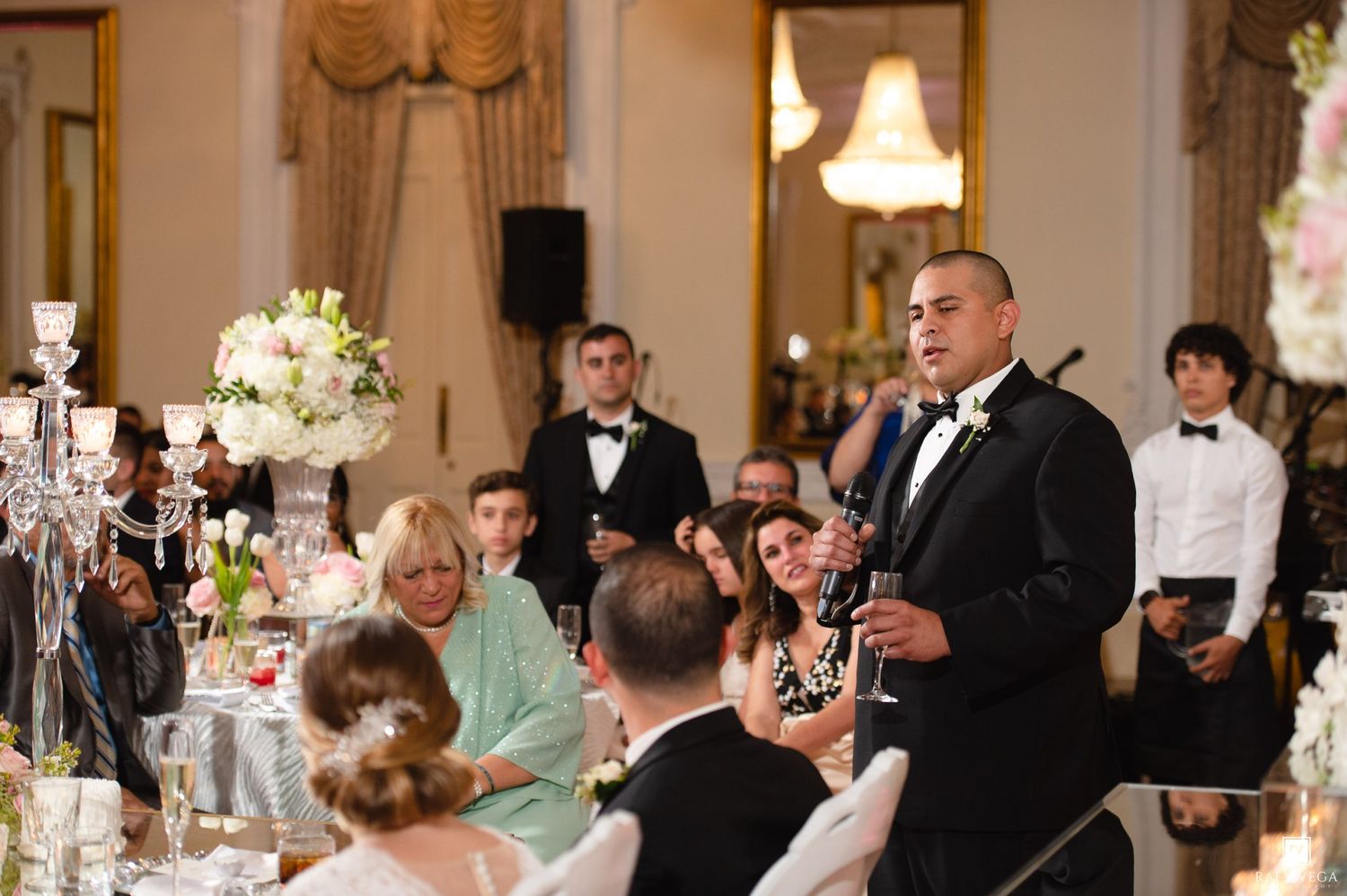 Man in tuxedo giving a speech at an elegant wedding reception with guests seated at decorated tables.