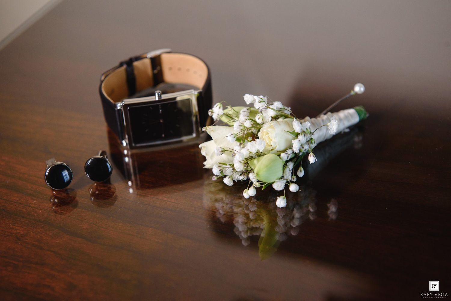Groom wedding accessories on wooden table featuring black leather watch, dark cufflinks, and white floral boutonniere.