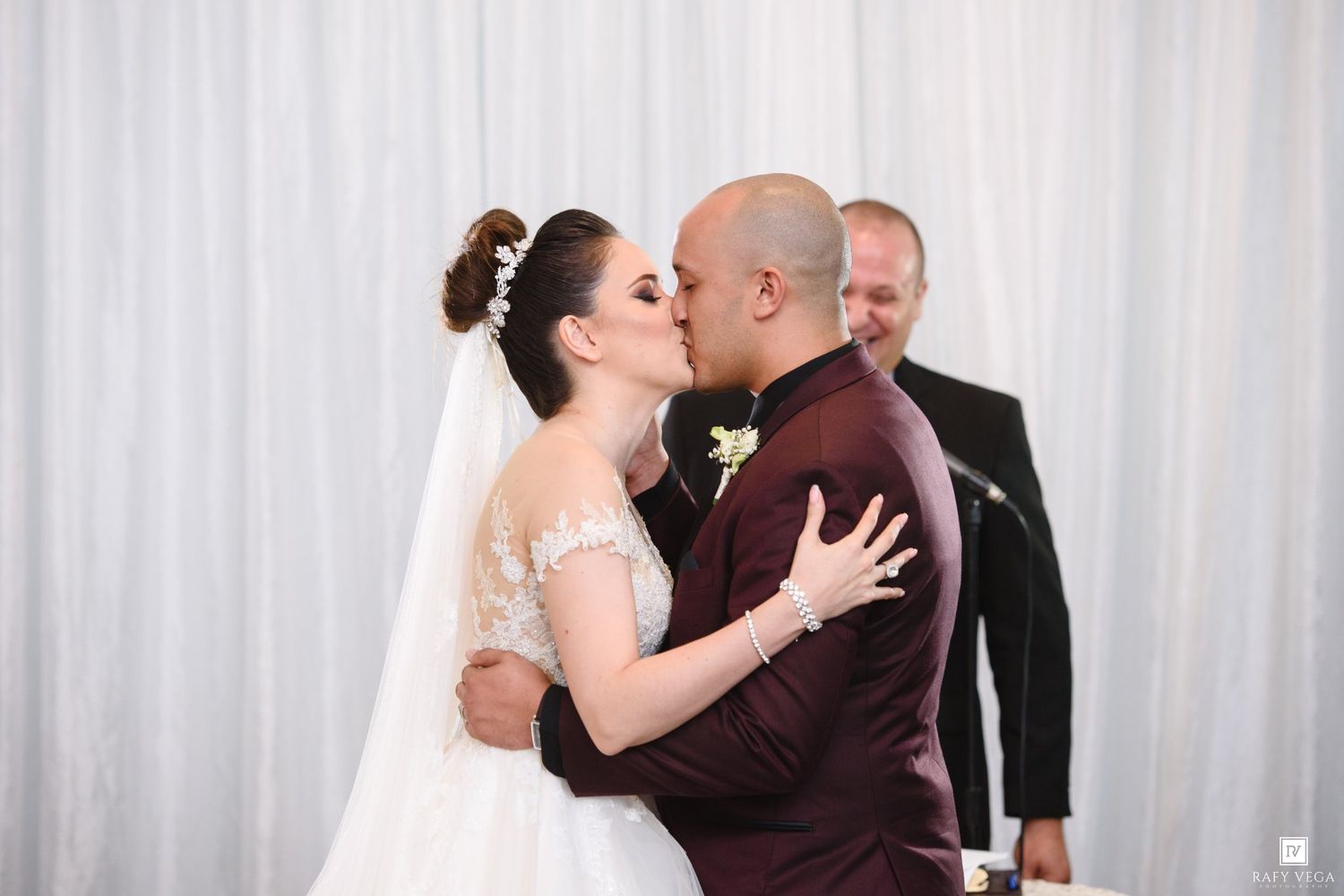 Bride and groom share first kiss at wedding altar, officiant standing behind them, bride in lace gown.