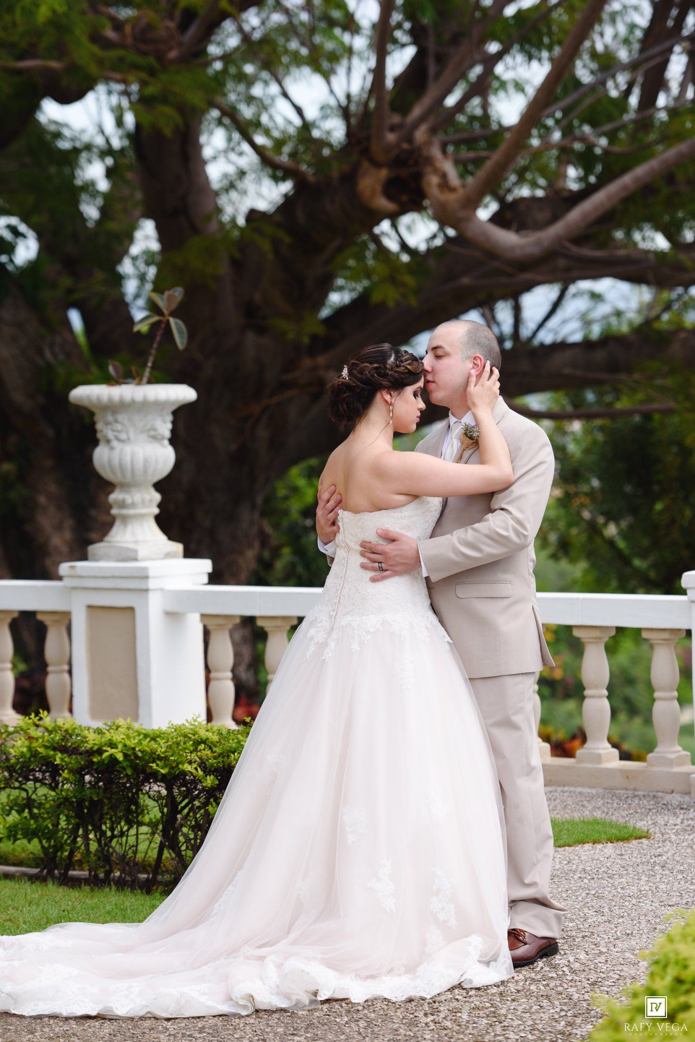 Bride and groom share a romantic kiss on an elegant white balcony surrounded by lush greenery on their wedding day.
