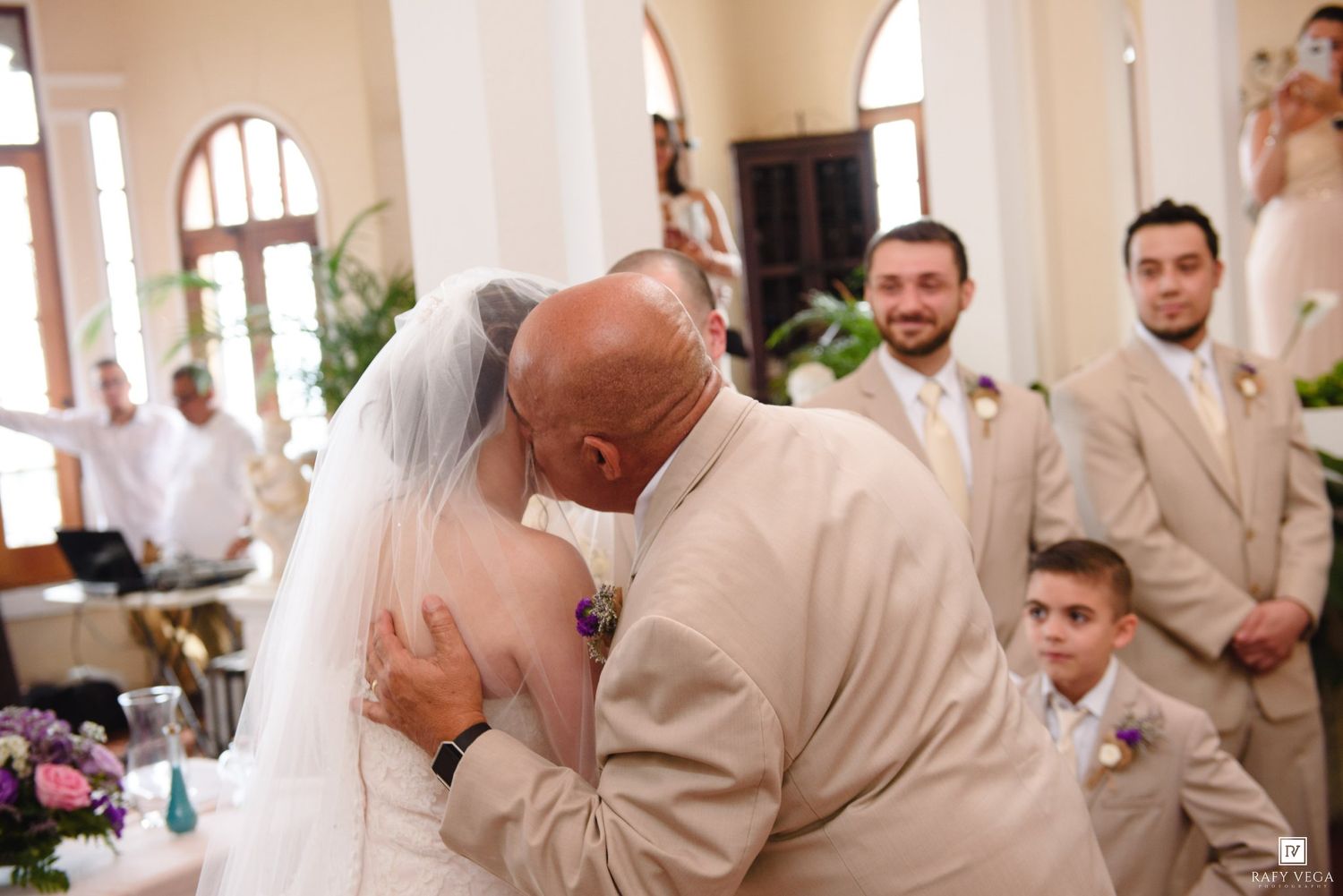 Bride and groom sharing a kiss at wedding ceremony with groomsmen and young ring bearer watching in elegant venue.
