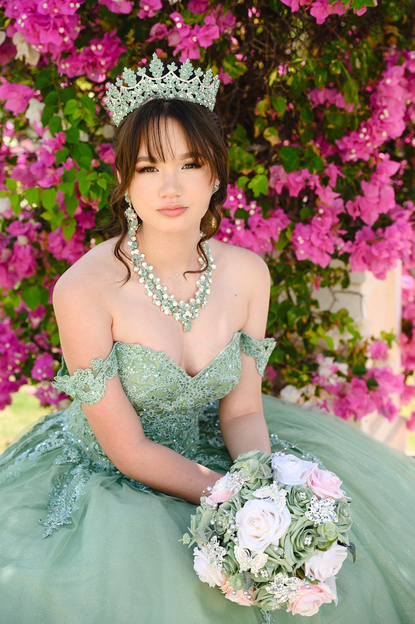Young woman in green quinceañera gown with tiara and bouquet poses before vibrant pink bougainvillea blooms.