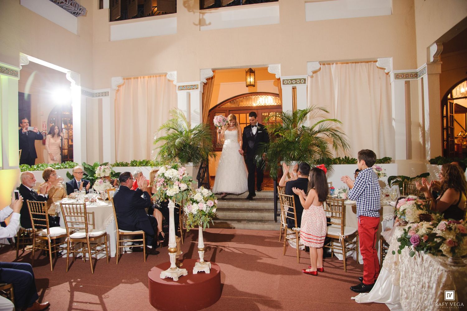 Elegant indoor wedding ceremony with bride and groom at altar, floral arrangements, gold chairs, and guests in a grand ballroom.