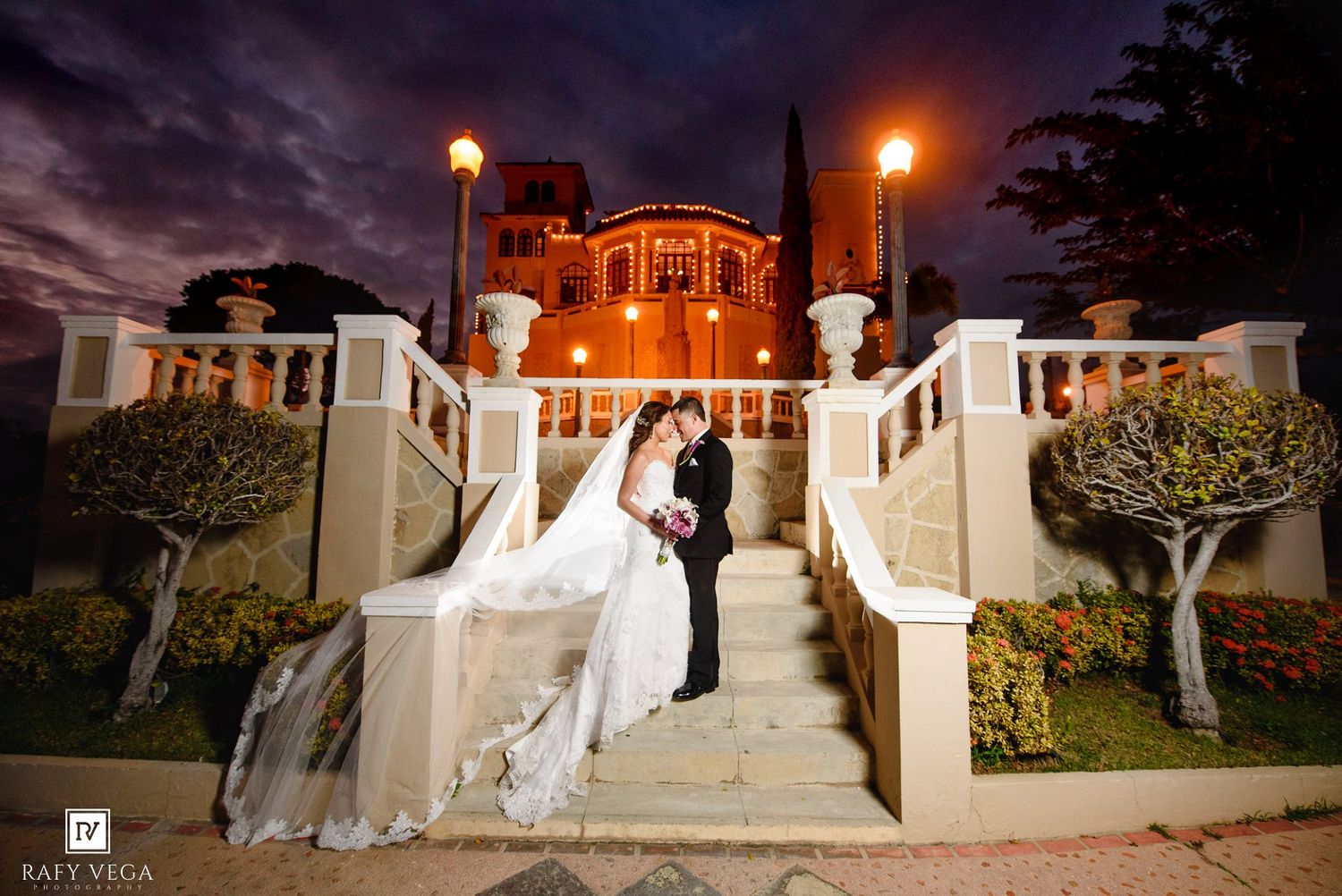 Bride and groom share romantic kiss on grand staircase outside illuminated historic venue at night.
