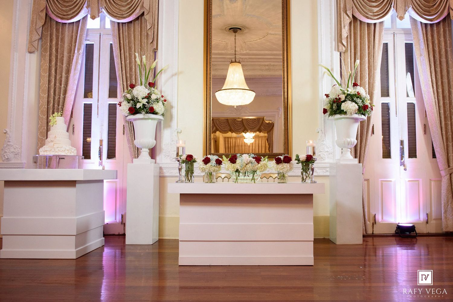 Elegant wedding reception venue with white floral arrangements, tall urns, a bar table, and warm uplighting against ornate windows.