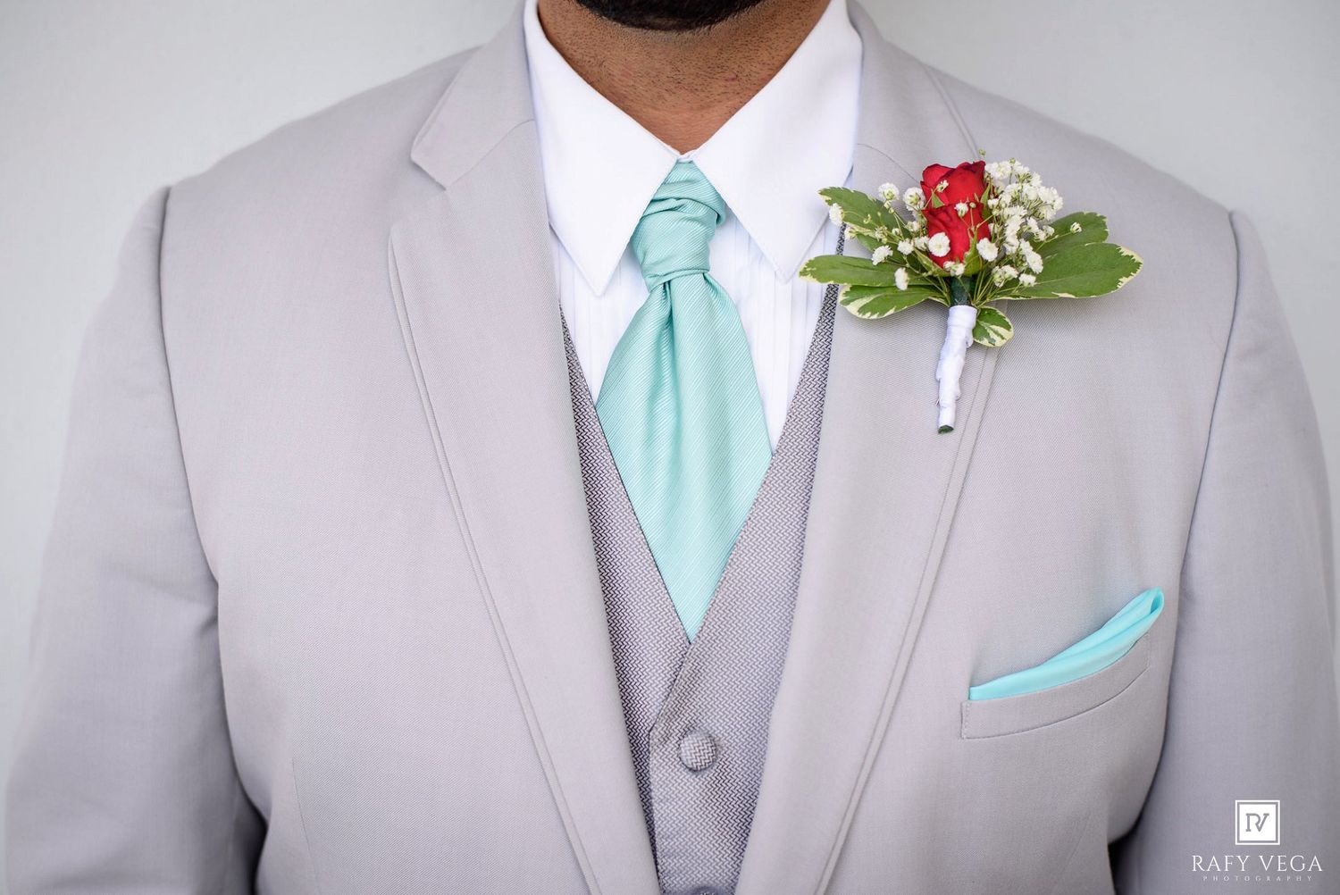 Groom in gray suit with mint green tie, matching pocket square, and red floral boutonniere with white baby's breath.