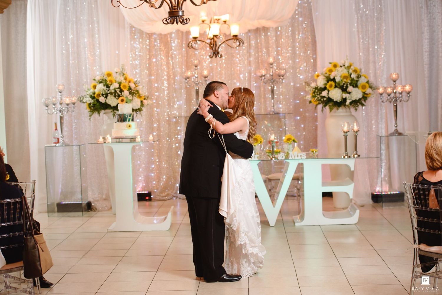 Bride and groom share first dance kiss in elegant ballroom with LOVE letters, floral arrangements, and twinkling backdrop lights.