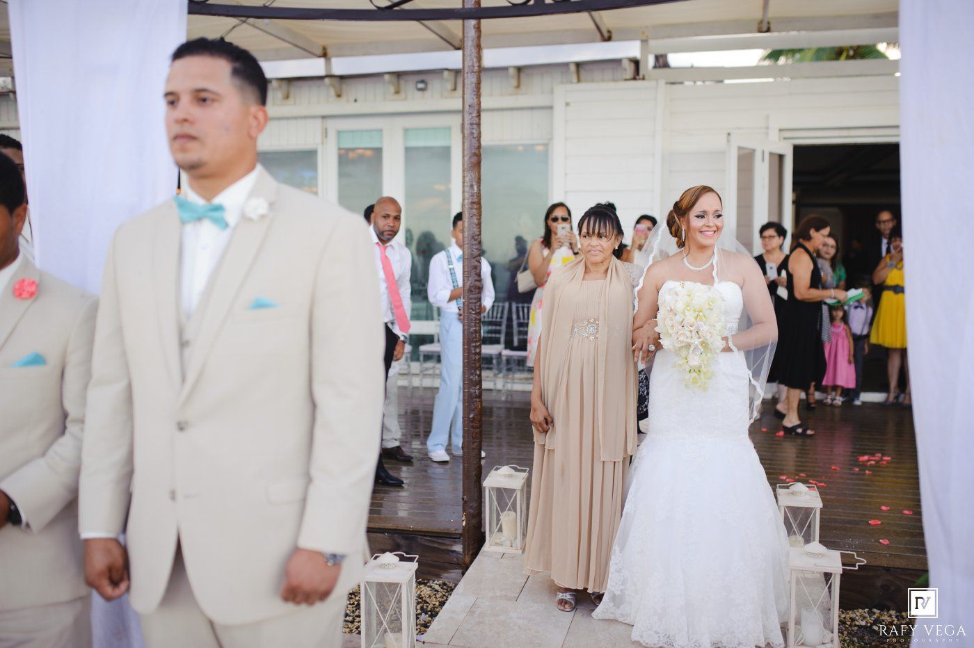 Bride in white mermaid gown walks down aisle with escort at outdoor wedding ceremony, groom in tan suit waits ahead.