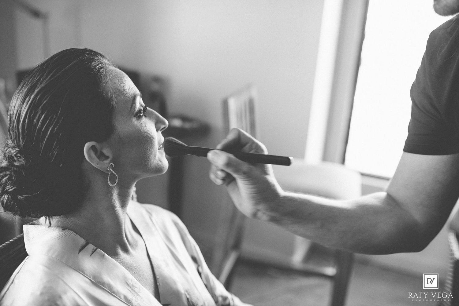 Black and white photo of a bride getting makeup applied with a brush by a makeup artist before her wedding.
