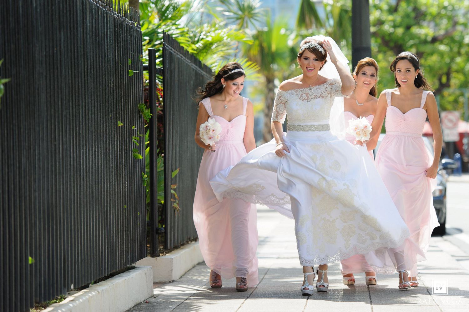 Bride in white gown walks with three bridesmaids in pink dresses along a sunny sidewalk on her wedding day.