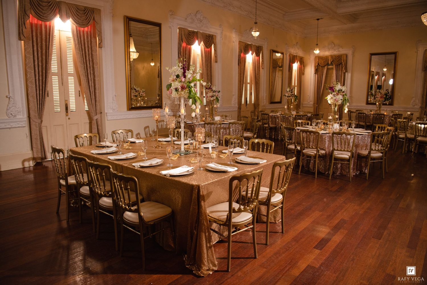 Elegant wedding reception hall with gold tablecloths, chiavari chairs, tall floral centerpieces, and hardwood floors in a classic ballroom.