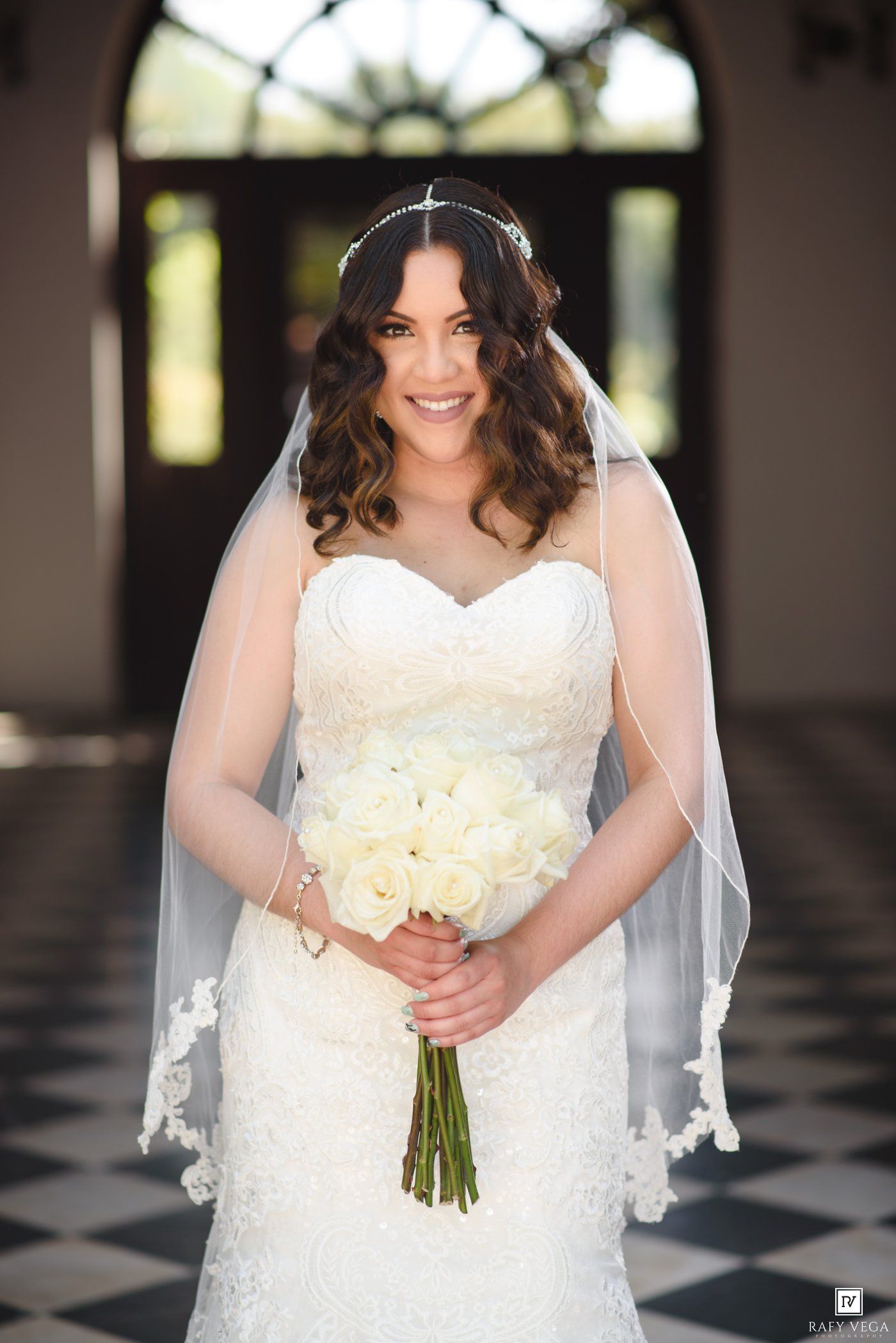Smiling bride in lace strapless gown holding white rose bouquet with veil and headpiece in elegant venue.