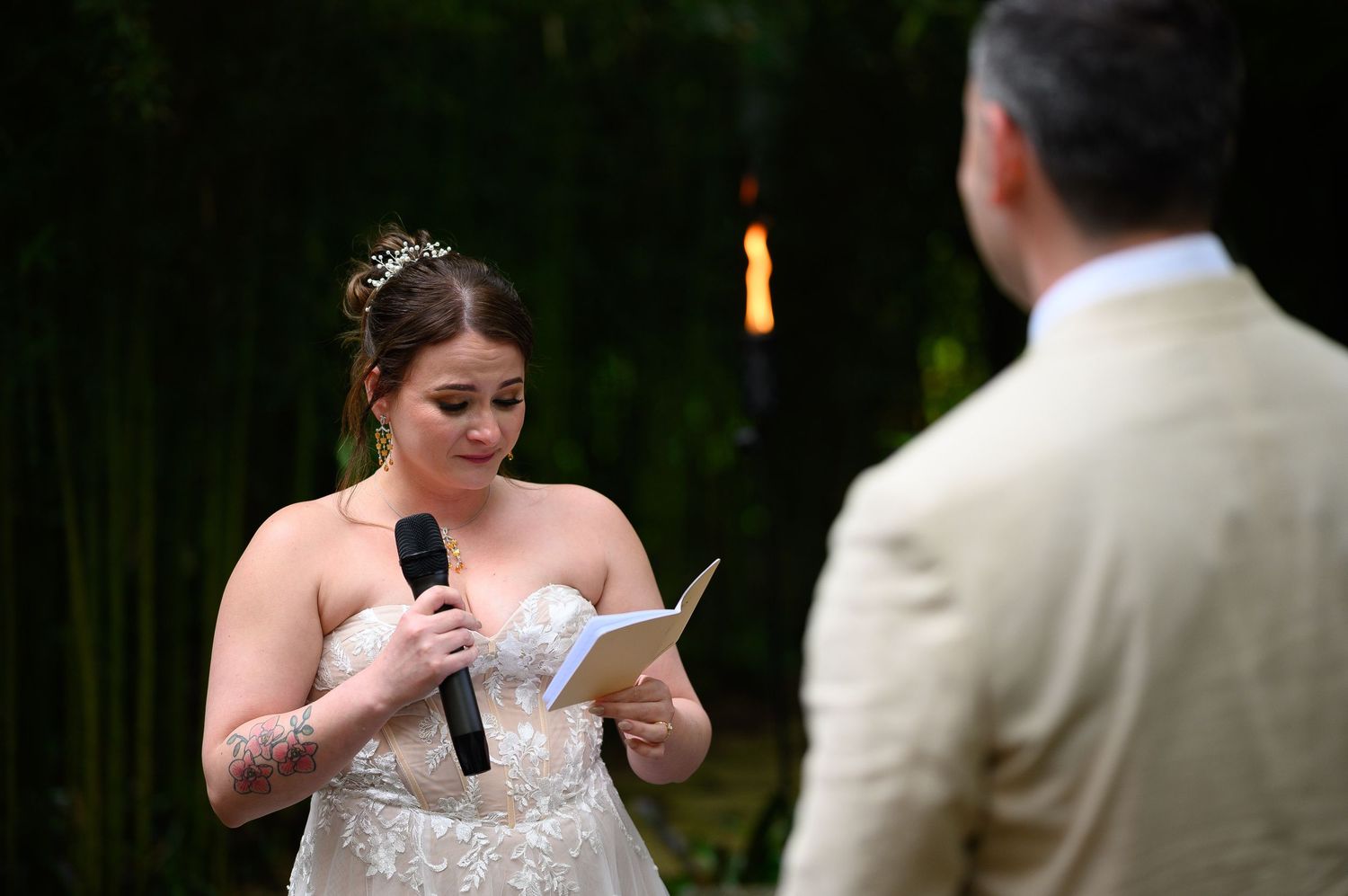 Bride reading wedding vows from a booklet while holding a microphone during an outdoor ceremony.