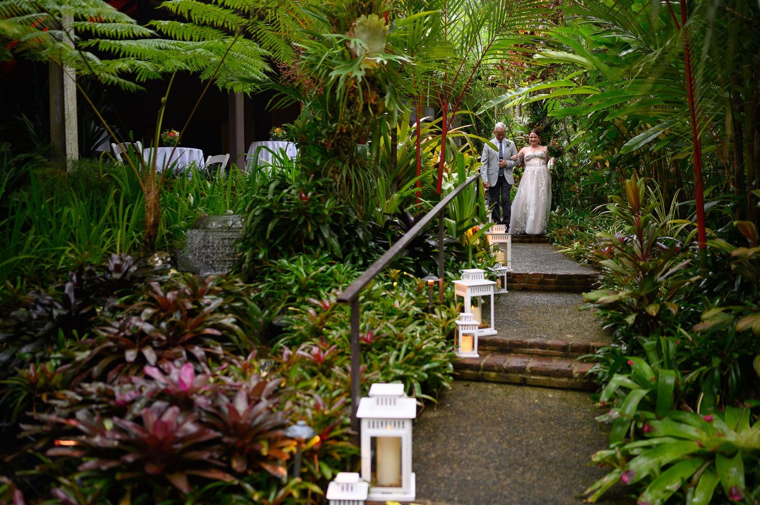 Couple in wedding attire walking along a lantern-lined garden path surrounded by lush tropical greenery.