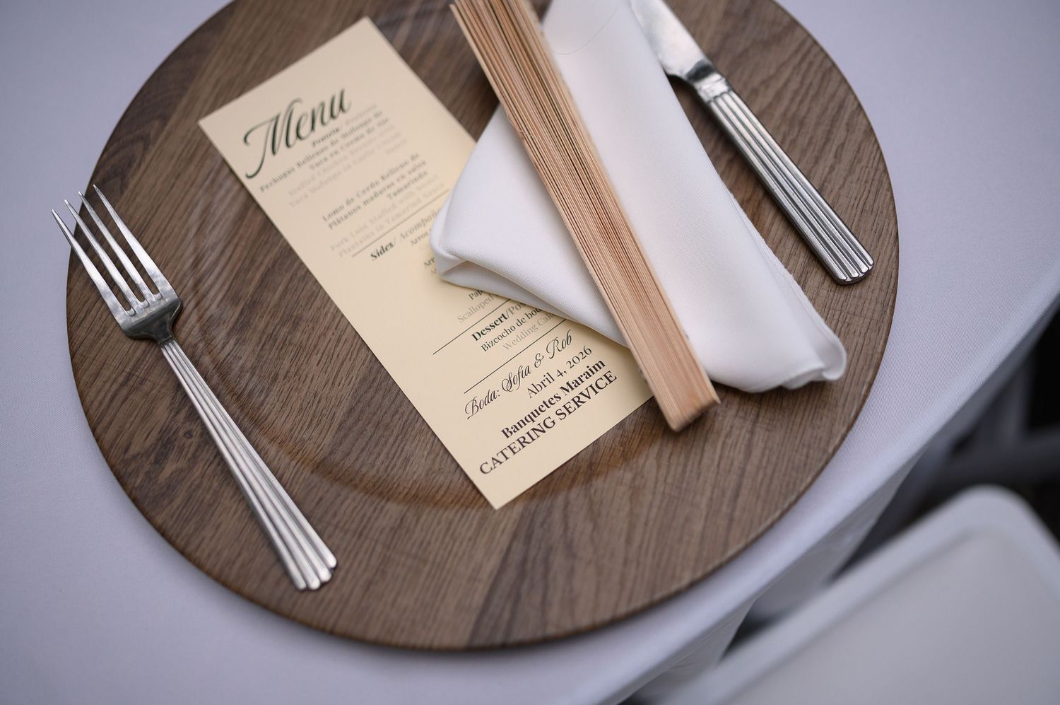 Elegant place setting with wooden charger plate, silverware, white napkin, and a catering service menu card.