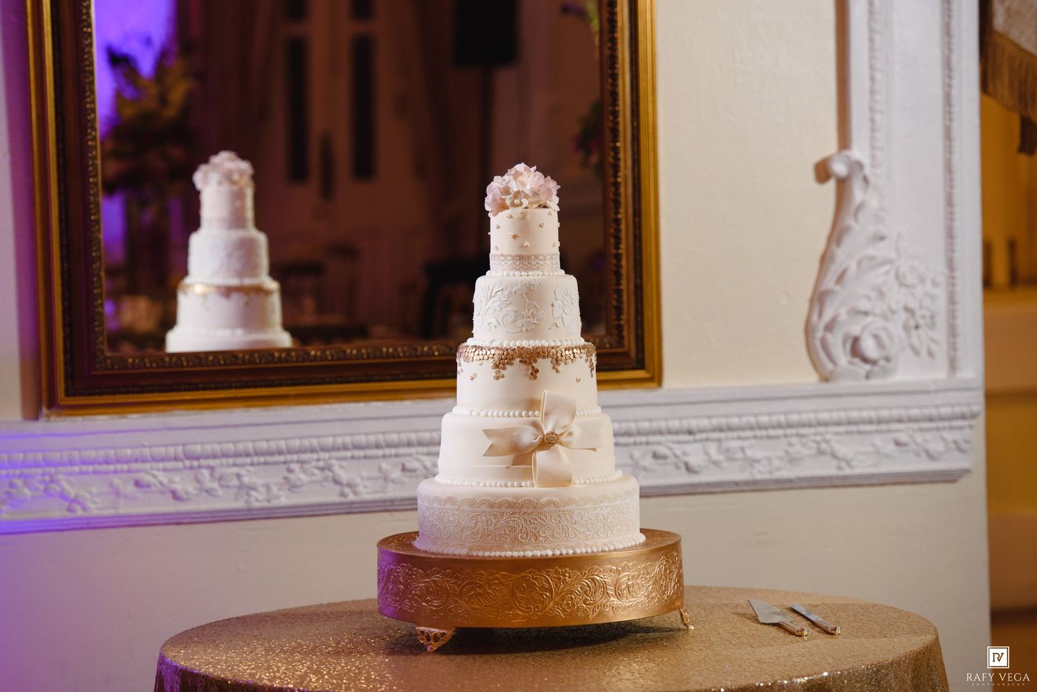 Elegant five-tier white wedding cake with gold accents displayed on a gold stand at a wedding reception.