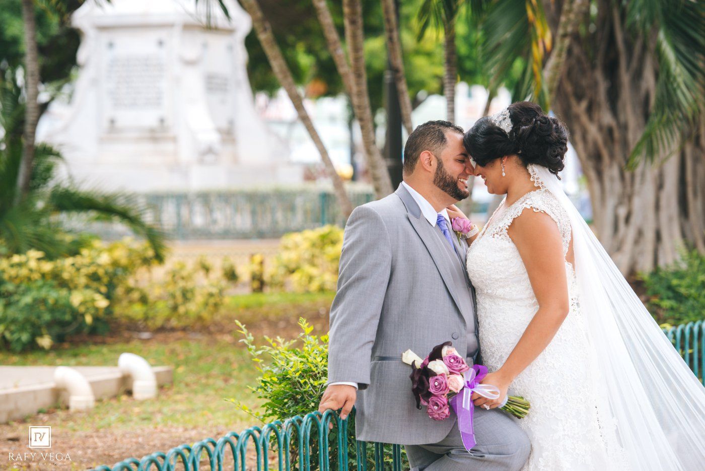 Bride and groom share romantic moment in park, bride holds purple bouquet, groom in gray suit at outdoor wedding.
