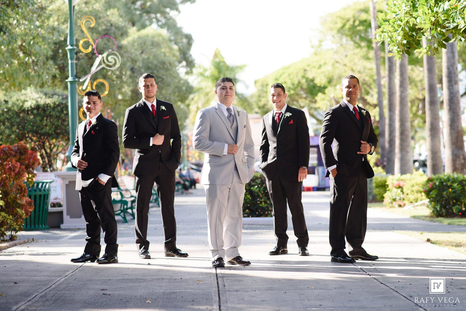 Groom in light gray suit poses with four groomsmen in black suits and red boutonnieres on a sunny outdoor walkway.