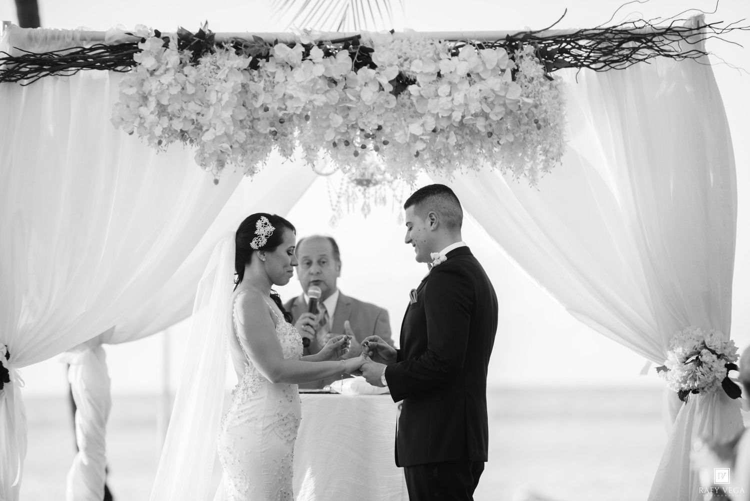 Black and white photo of a couple exchanging vows under a floral arch at an outdoor beach wedding ceremony.