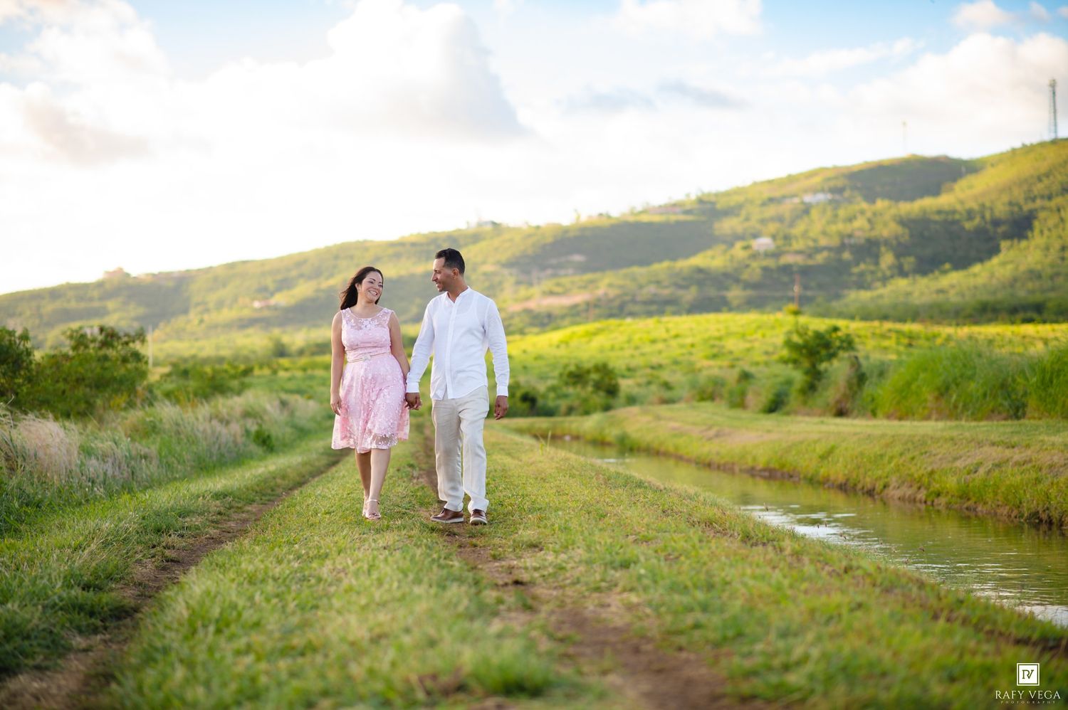 Couple holding hands walking on a scenic green path with rolling hills and a stream in the background.