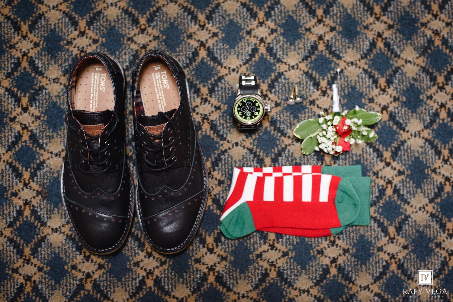Black wingtip dress shoes, festive red and green striped socks, a watch, and floral boutonniere on a plaid surface.
