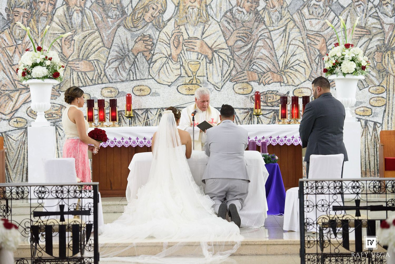 A bride and groom kneel at the altar during a church wedding ceremony with a priest officiating before a mosaic backdrop.