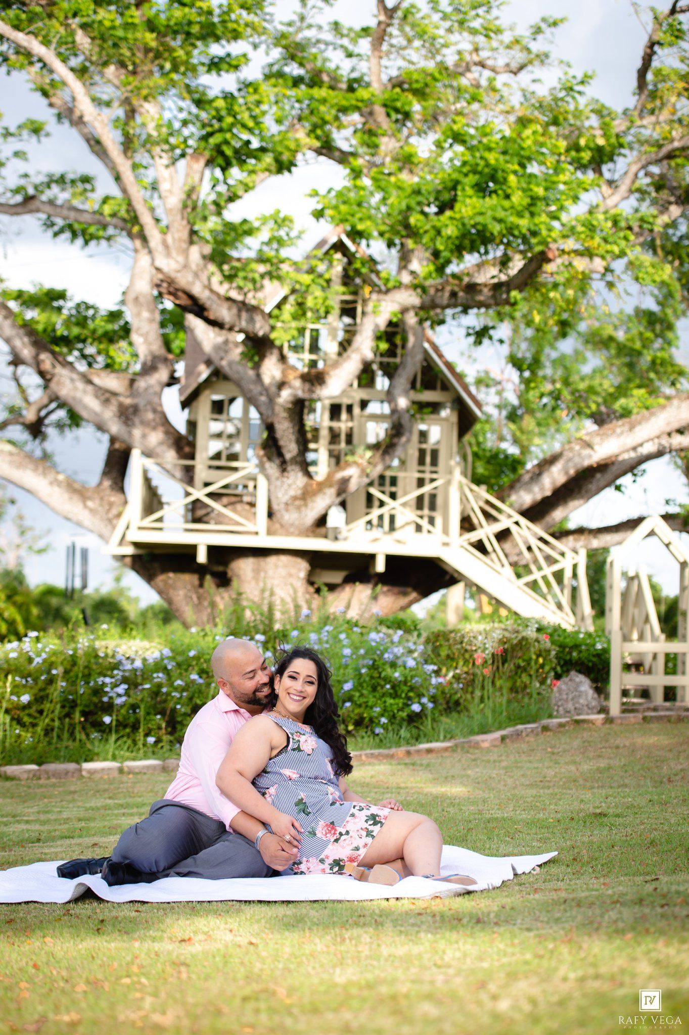 Couple sitting on white blanket in garden with treehouse backdrop for romantic outdoor maternity photo session.