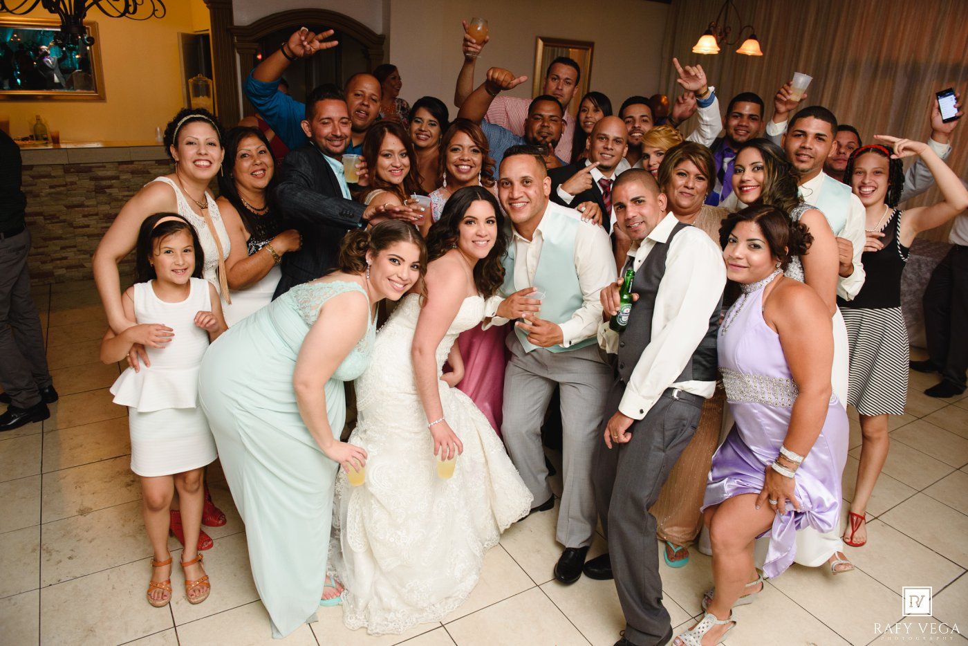 A joyful wedding reception group photo with bride and groom surrounded by smiling guests in elegant attire.