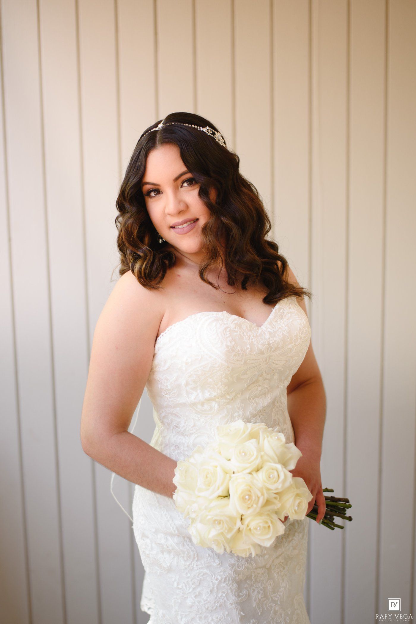 Bride in strapless lace mermaid gown holding white rose bouquet with tiara and curled hair.