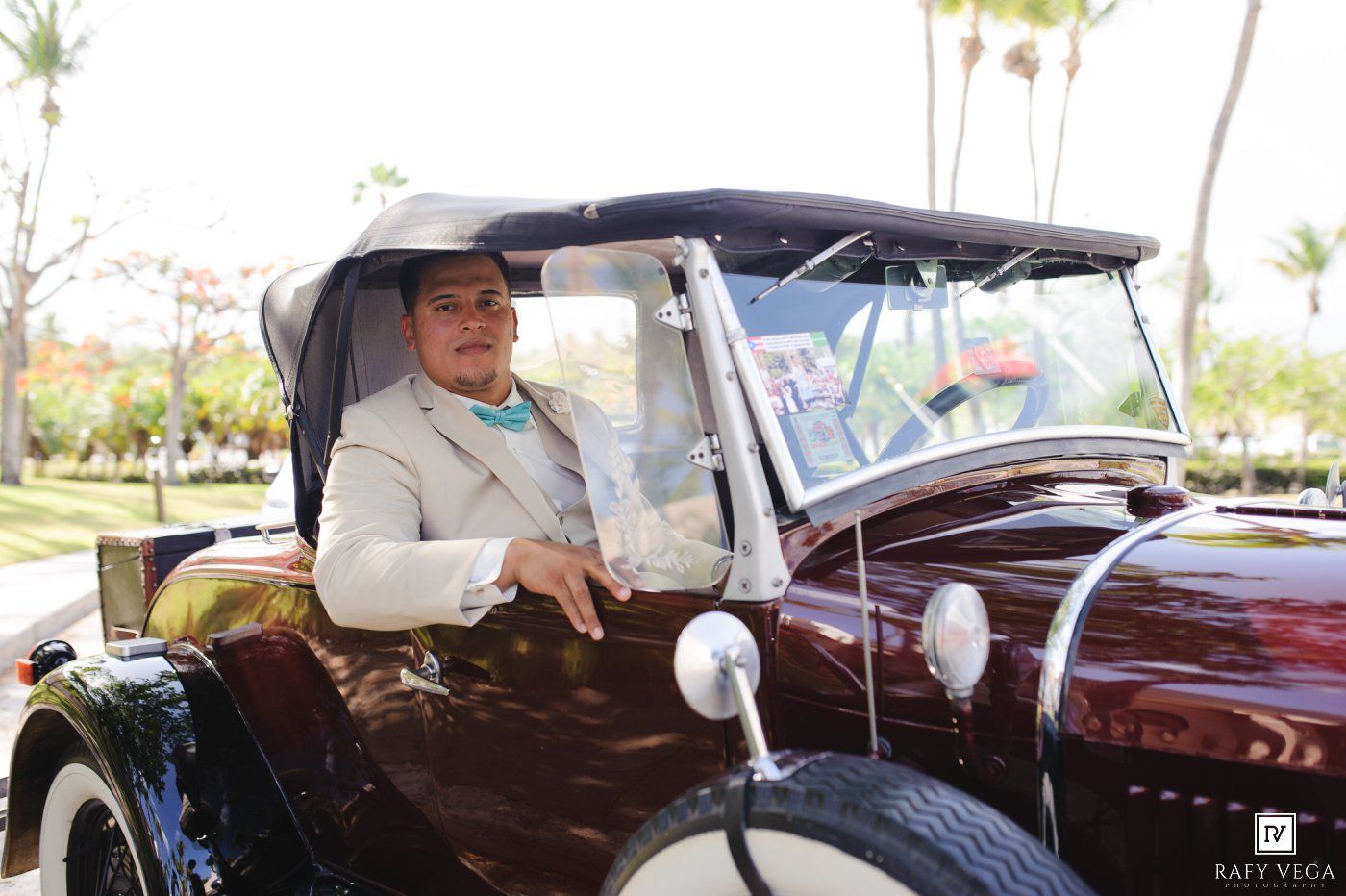 Elegant man in beige suit driving a vintage classic convertible car at a tropical venue.