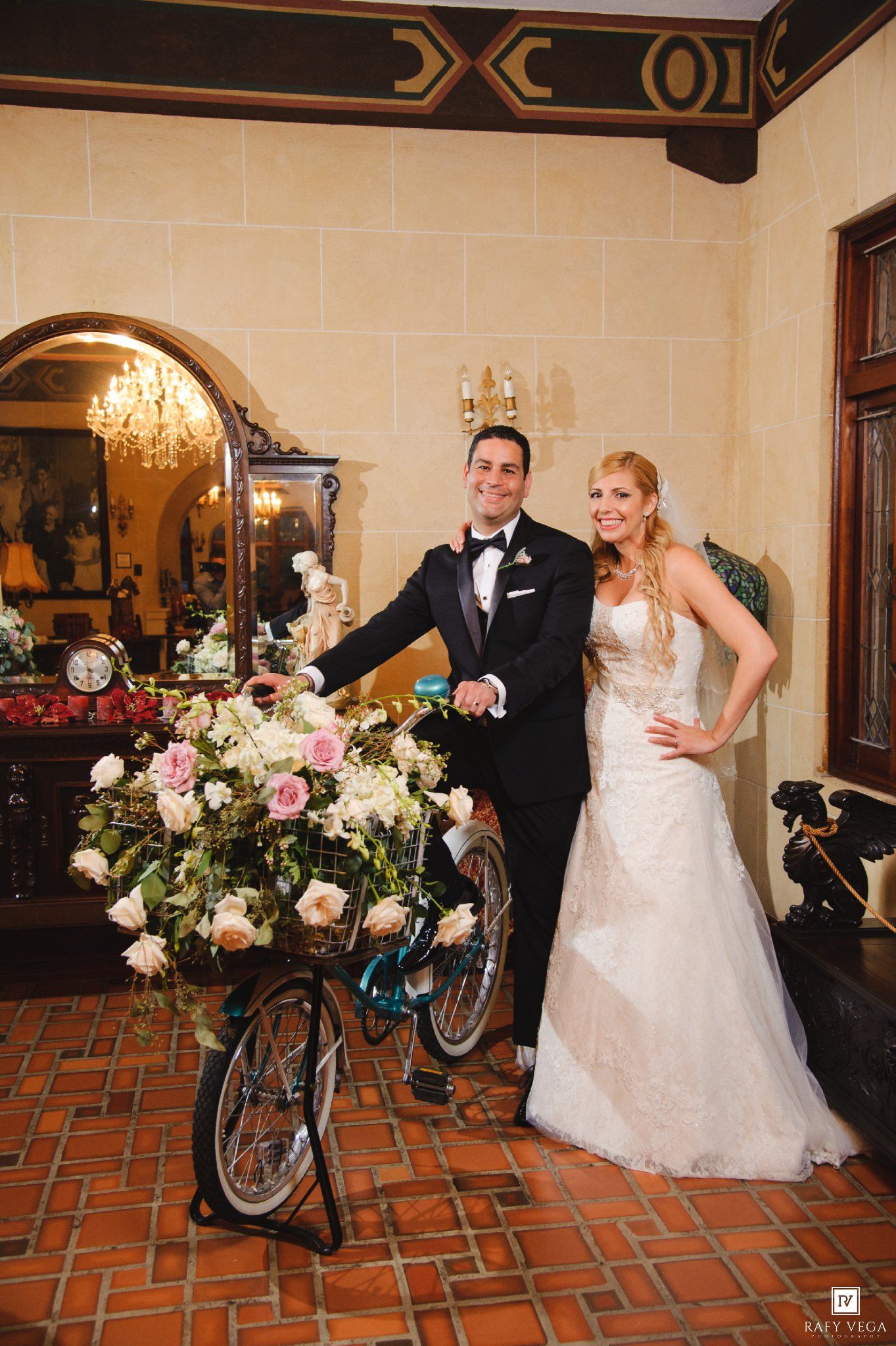 Bride and groom posing with a flower-adorned bicycle inside an elegant venue with arched mirrors and chandelier.
