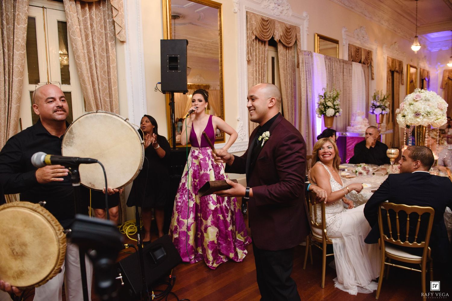 Elegant wedding reception with live drummers, guests in formal attire, floral centerpieces, and a woman in floral gown.