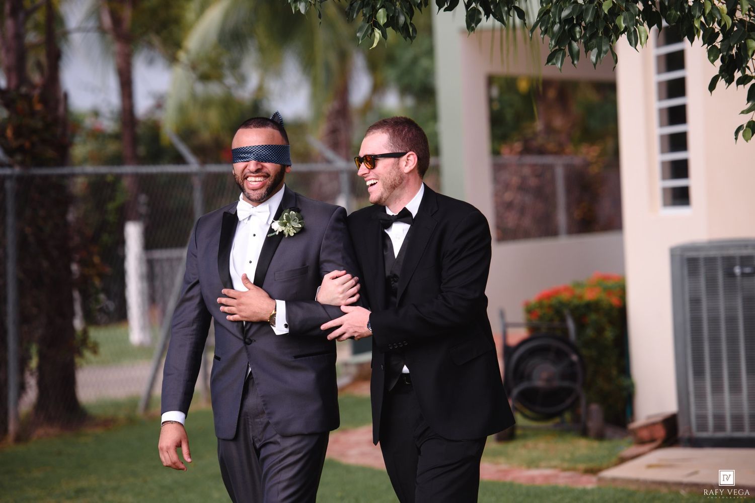 Two groomsmen in suits walk arm in arm outdoors, one wearing a blindfold, at a wedding celebration.