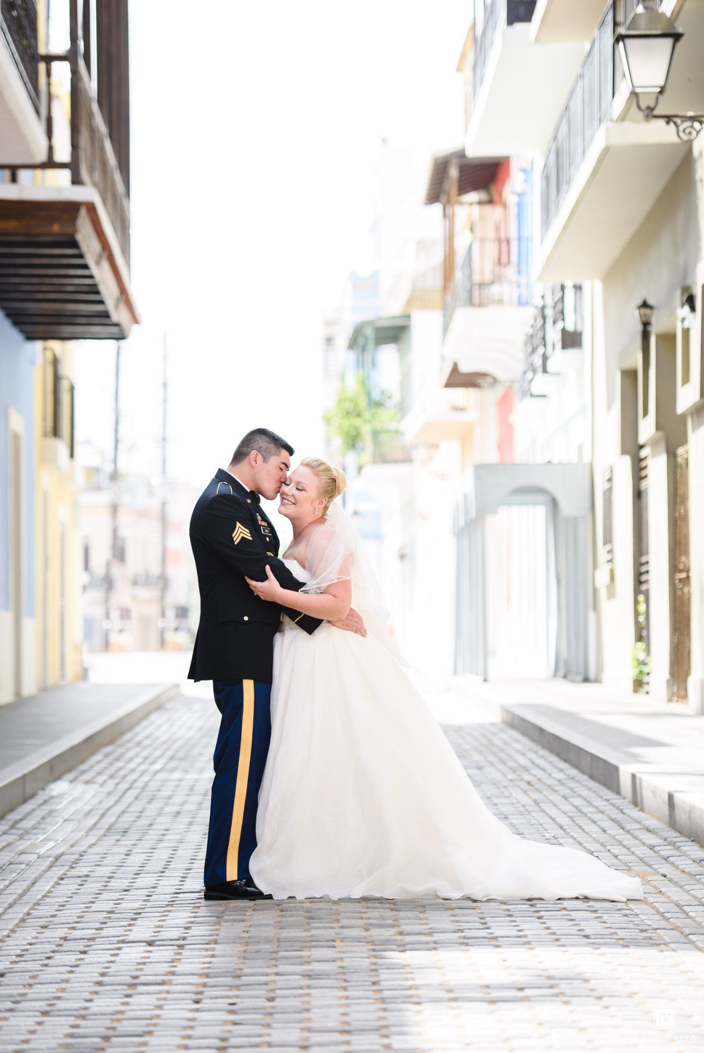 Military groom in dress uniform embraces bride in white gown on cobblestone street in Old San Juan, Puerto Rico.
