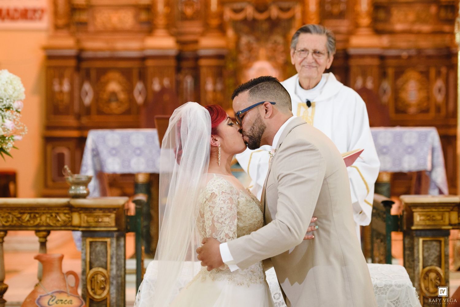 Bride and groom share first kiss at church altar as smiling priest watches during wedding ceremony.