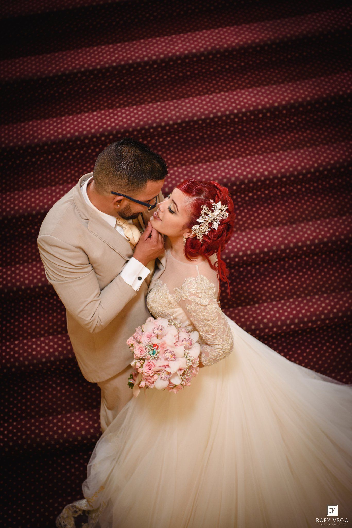 Bride with red hair holding pink bouquet shares romantic kiss with groom in beige suit against burgundy backdrop.