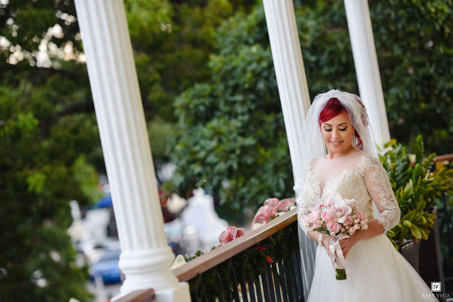 Bride in lace wedding dress holding pink bouquet stands on elegant white columned balcony surrounded by lush greenery.