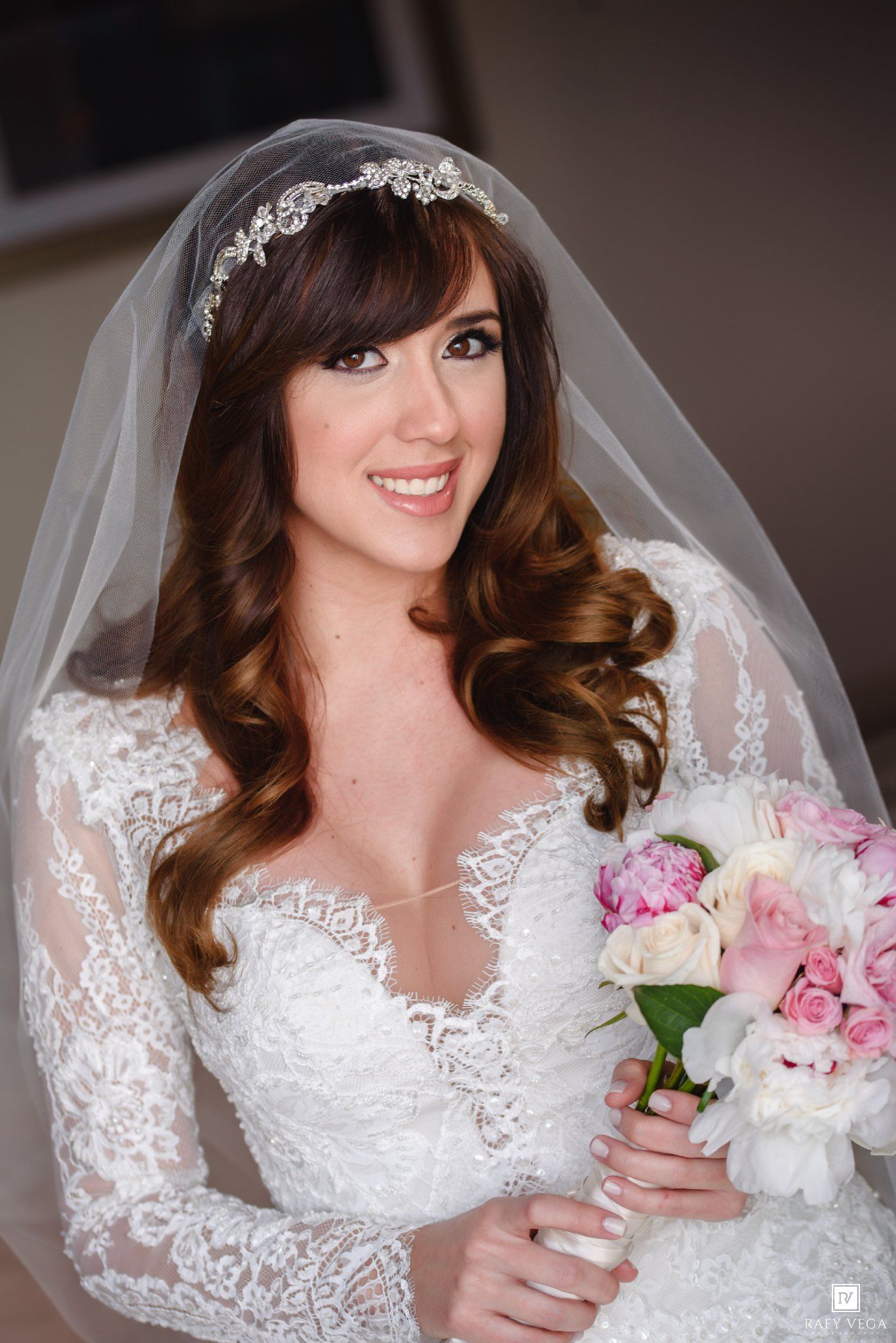 Smiling bride in lace long-sleeve gown and veil headpiece holds pink and white floral bouquet.