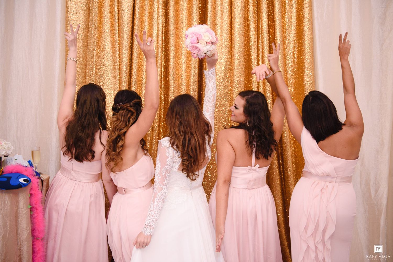 Bride and bridesmaids in pink dresses facing gold sequin backdrop, arms raised as bouquet is tossed at wedding reception.