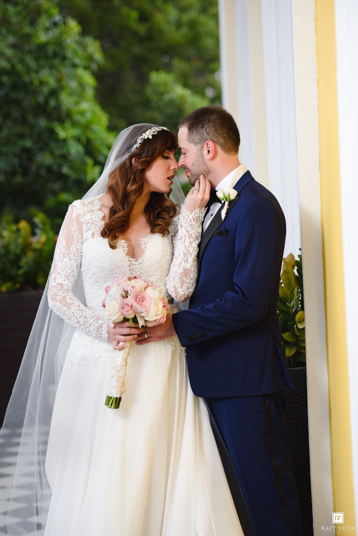 Bride in lace long-sleeve gown with veil tenderly touches grooms chin while holding pink roses bouquet.