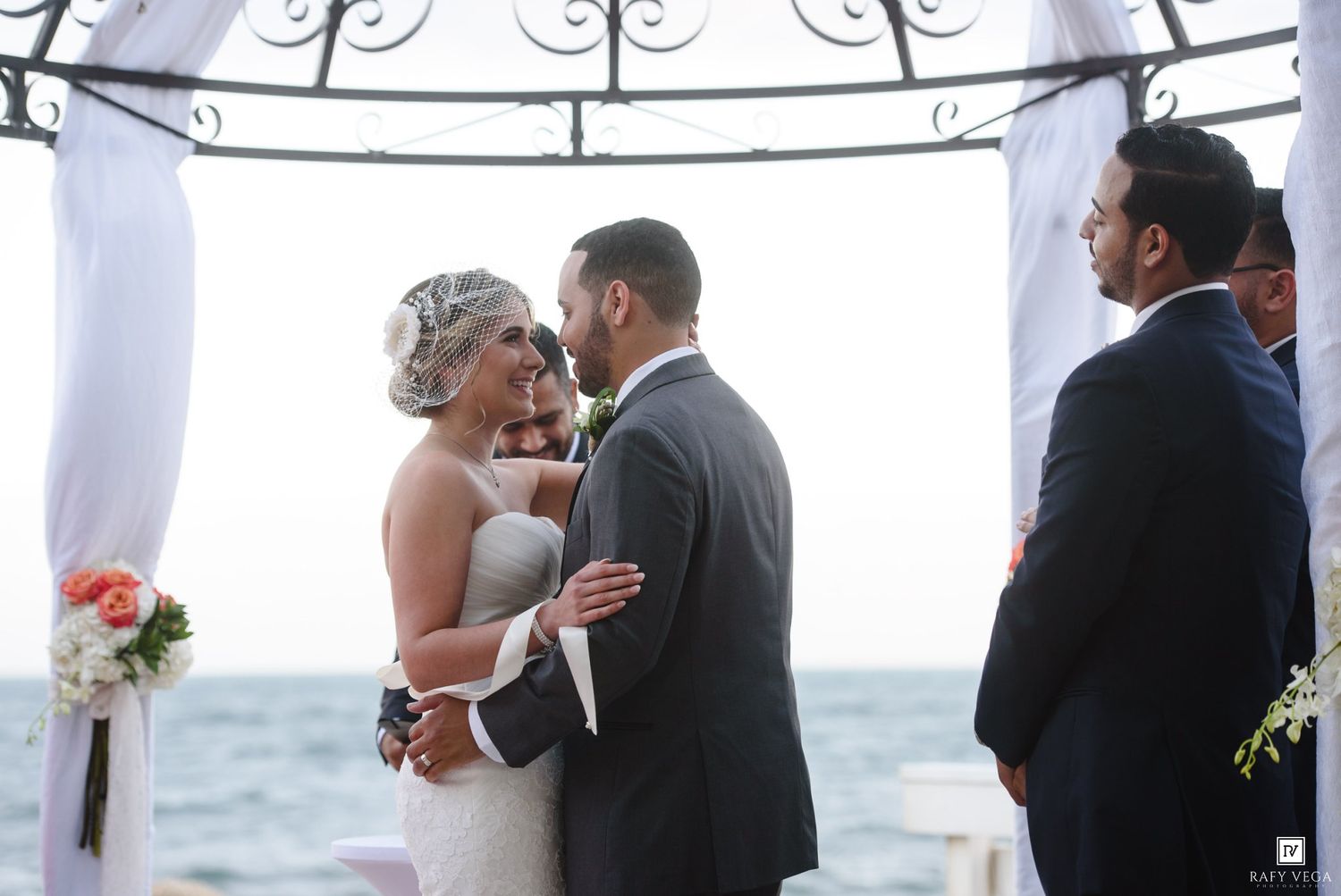 Bride and groom share a romantic moment at an outdoor oceanfront wedding ceremony under a white draped arch.