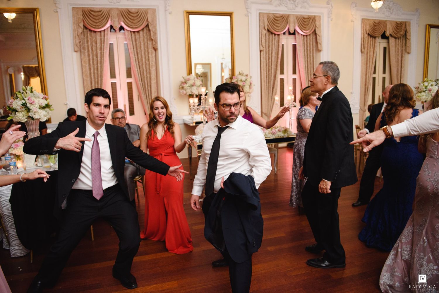 Men dancing energetically at an elegant wedding reception in a decorated ballroom with floral arrangements.