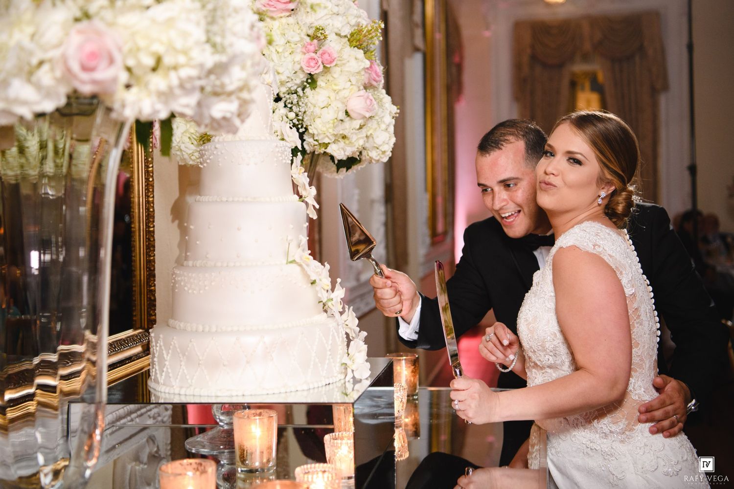 Bride and groom cut their elegant white wedding cake surrounded by candles and lush floral centerpieces.