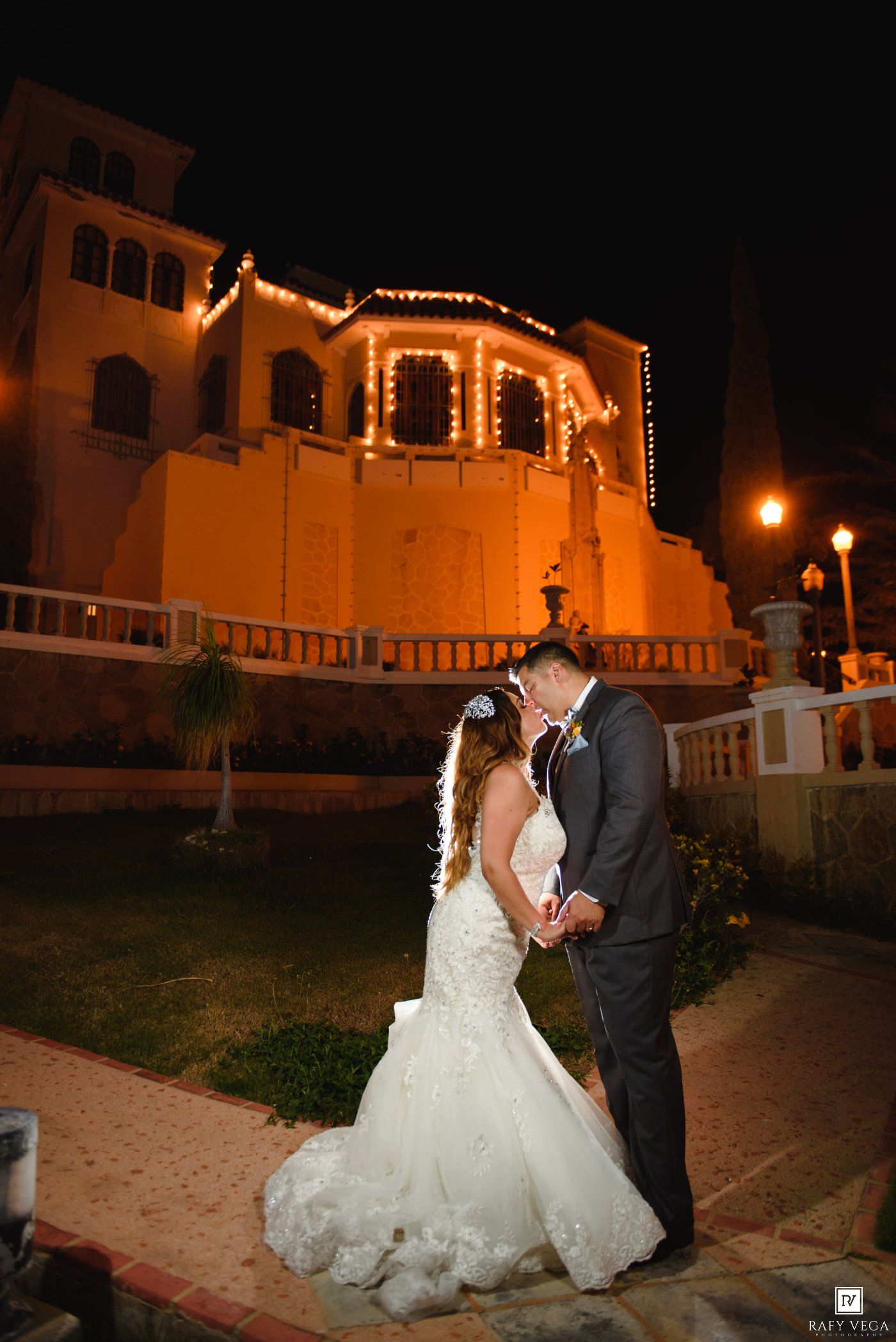 Bride and groom share a romantic moment outside an illuminated venue at night, bride in a lace mermaid gown.
