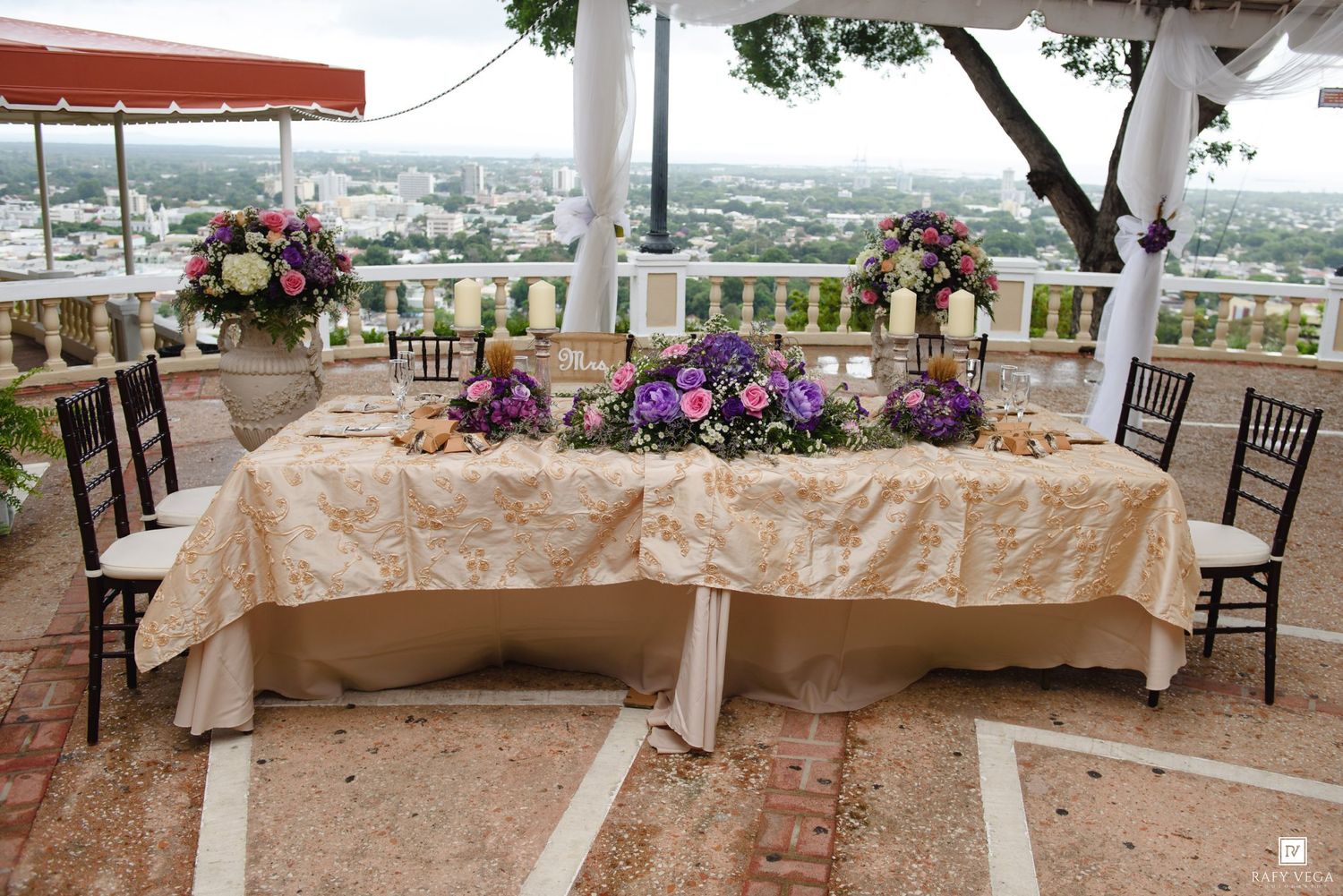 Elegant outdoor wedding reception table with purple floral centerpieces and gold tablecloth.
