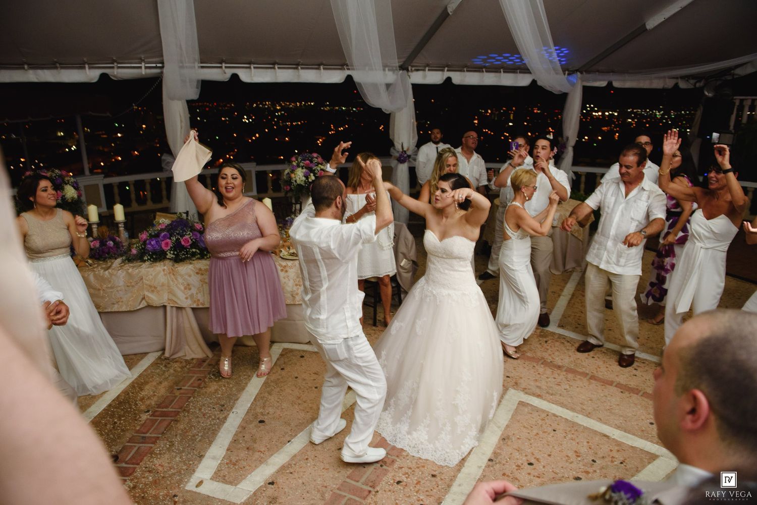 Bride and groom dancing joyfully with guests at an elegant outdoor wedding reception at night.